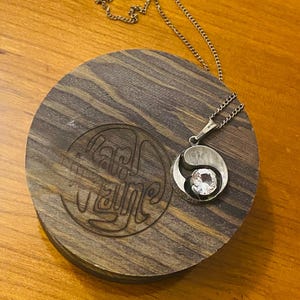May include: A silver necklace with a yin yang pendant featuring a clear crystal. The necklace is displayed on a wooden box with the text "Pam Wayne" engraved on the lid.
