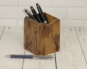 May include: A wooden pen holder with a unique tree-like design, holding several pens. A pen with a blue patterned barrel rests in front of the holder. The holder and pen are on a light wood surface.