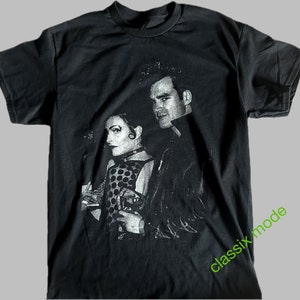 May include: Black t-shirt with a silver print of a man and woman. The man is wearing a leather jacket and the woman is wearing a polka dot dress. The text "classix mode" is printed in green on the bottom right corner of the shirt.
