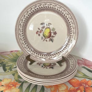 May include: A set of vintage ceramic plates with a pear and floral design. The plates have a cream-colored center with a brown border featuring a repeating leaf pattern. The pear is yellow and green with pink and green floral accents.