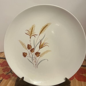 May include: A round white ceramic dinner plate featuring a central botanical illustration of wheat stalks and autumn-coloured leaves in shades of golden yellow, brown, and muted blue. The plate has a smooth, glossy finish and a simple, clean edge.