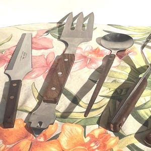 May include: A collection of vintage kitchen utensils with stainless steel heads and wooden handles. The set includes a slotted spoon, pie server, fork, spoon, masher, and a knife. The utensils are arranged on a floral patterned cloth.
