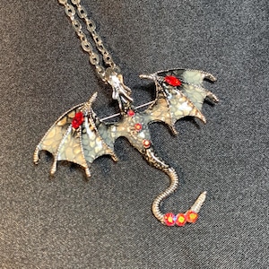 May include: A silver-toned dragon pendant necklace featuring wings with a textured, pale green and grey scale pattern. The dragon body is accented with small, round red crystals and larger, marquise-cut red stones on each wing.
