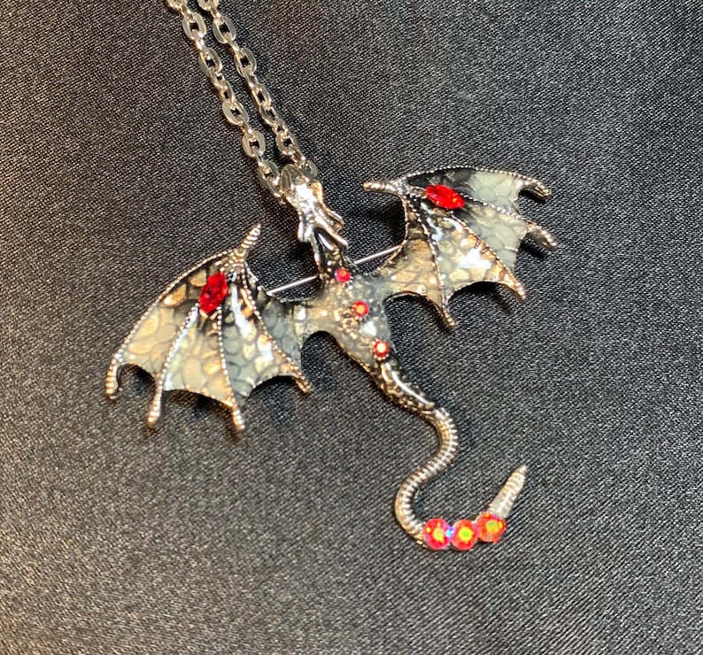 May include: A silver-toned dragon pendant necklace featuring wings with a textured, pale green and grey scale pattern. The dragon body is accented with small, round red crystals and larger, marquise-cut red stones on each wing.