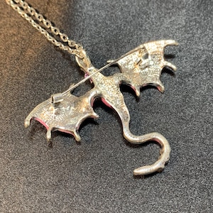 May include: A silver-toned metal dragon pendant necklace featuring a textured finish on the reverse side. The dragon design includes spread wings and a long, curved tail. A horizontal pin mechanism is attached to the back of the dragon figure.