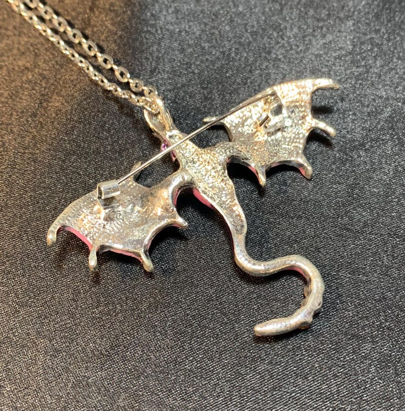 May include: A silver-toned metal dragon pendant necklace featuring a textured finish on the reverse side. The dragon design includes spread wings and a long, curved tail. A horizontal pin mechanism is attached to the back of the dragon figure.