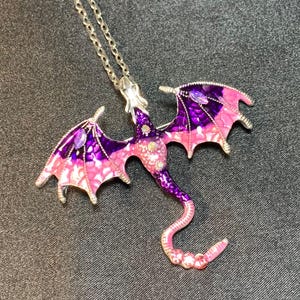 May include: A silver-toned metal dragon pendant necklace featuring enamel detailing in shades of purple and pink. The dragon has outstretched wings with purple and pink gradients, a long curved tail, and small embedded clear crystals on its body.