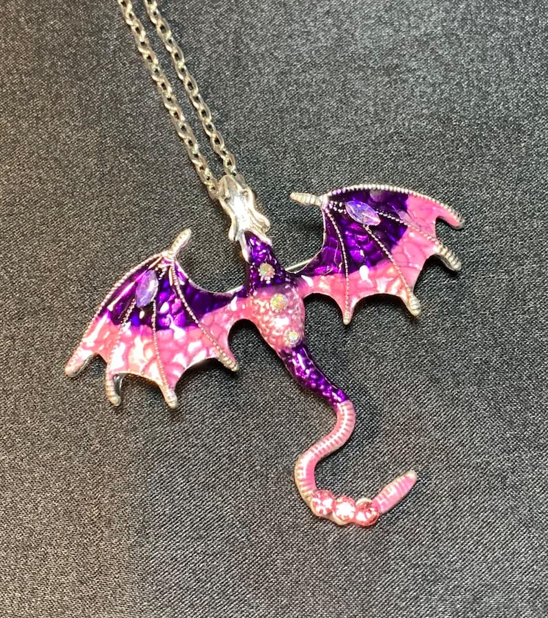 May include: A silver-toned metal dragon pendant necklace featuring enamel detailing in shades of purple and pink. The dragon has outstretched wings with purple and pink gradients, a long curved tail, and small embedded clear crystals on its body.