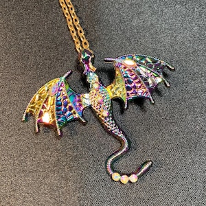 May include: A dragon-shaped pendant necklace featuring an iridescent, multi-colored metallic finish with purple, blue, and gold tones. The dragon body and wings are textured with a scale-like pattern and accented with small, clear, reflective rhinestones.