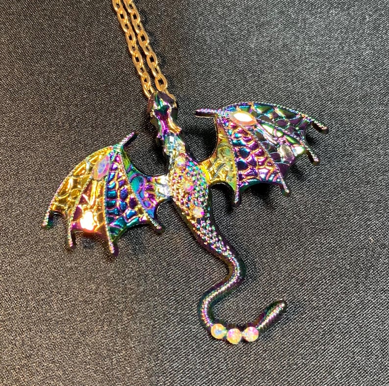 May include: A dragon-shaped pendant necklace featuring an iridescent, multi-colored metallic finish with purple, blue, and gold tones. The dragon body and wings are textured with a scale-like pattern and accented with small, clear, reflective rhinestones.