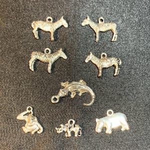 May include: A collection of seven silver-toned metal charms featuring animal shapes. The set includes two zebras, one alligator, one monkey, one rhinoceros, and one hippopotamus, each with a small circular loop at the top for attachment.