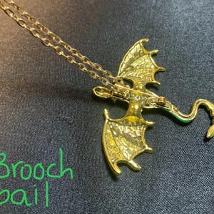 May include: A gold-toned dragon pendant necklace featuring a textured wing design and a curved tail. The back of the dragon includes a brooch bail mechanism, allowing the piece to function as both a pendant and a brooch. The text Brooch bail is visible.