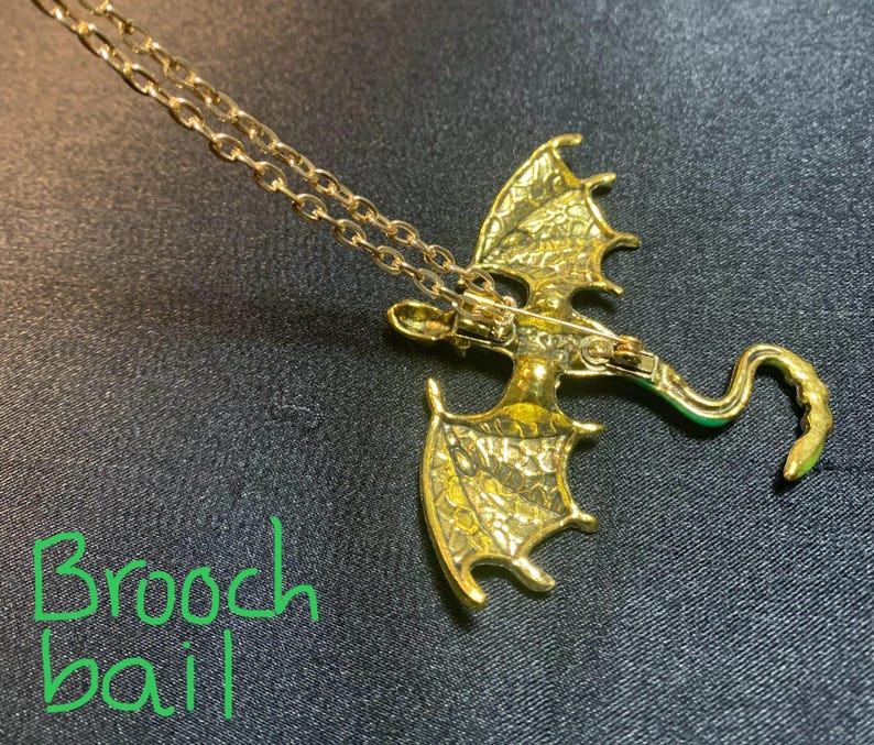 May include: A gold-toned dragon pendant necklace featuring a textured wing design and a curved tail. The back of the dragon includes a brooch bail mechanism, allowing the piece to function as both a pendant and a brooch. The text Brooch bail is visible.
