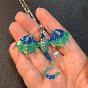 May include: A silver-toned dragon pendant necklace featuring enamel wings and body in gradient shades of blue and green. The dragon is embellished with small clear and blue rhinestones along its back, wings, and tail, suspended from a silver chain.