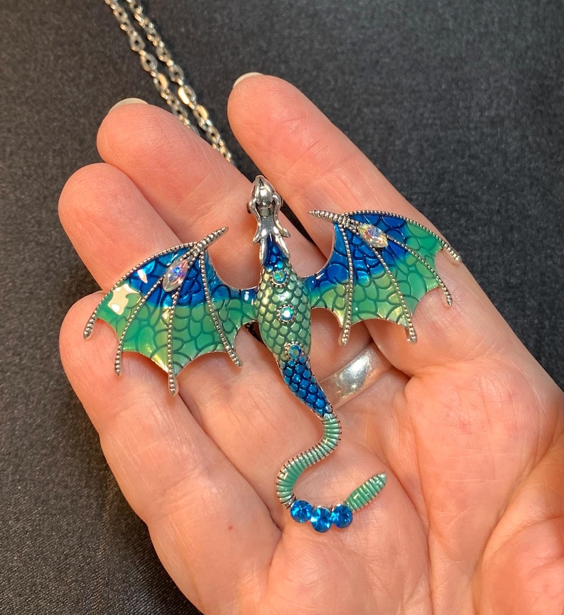 May include: A silver-toned dragon pendant necklace featuring enamel wings and body in gradient shades of blue and green. The dragon is embellished with small clear and blue rhinestones along its back, wings, and tail, suspended from a silver chain.