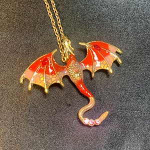 May include: A gold-toned dragon pendant necklace featuring vibrant red and orange enamel wings with gold scale detailing. The dragon body is adorned with small embedded crystals, including three pink stones at the tip of its curved tail.