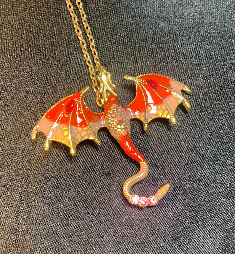 May include: A gold-toned dragon pendant necklace featuring vibrant red and orange enamel wings with gold scale detailing. The dragon body is adorned with small embedded crystals, including three pink stones at the tip of its curved tail.