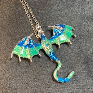 May include: A silver-toned metal dragon pendant necklace featuring an enamel finish in gradient shades of blue and green. The dragon wings and body are detailed with a scale pattern and small, sparkling inset crystals in blue and clear tones.