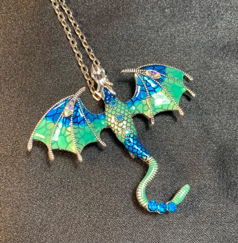 May include: A silver-toned metal dragon pendant necklace featuring an enamel finish in gradient shades of blue and green. The dragon wings and body are detailed with a scale pattern and small, sparkling inset crystals in blue and clear tones.