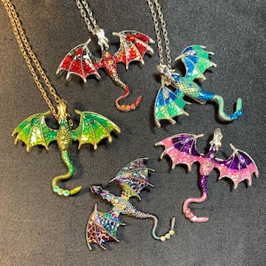 May include: Five dragon-shaped pendants on silver-toned chains. The dragons feature colorful enamel wings and bodies in red, green, blue, purple, and iridescent patterns, with small embedded crystals along their tails and bodies.