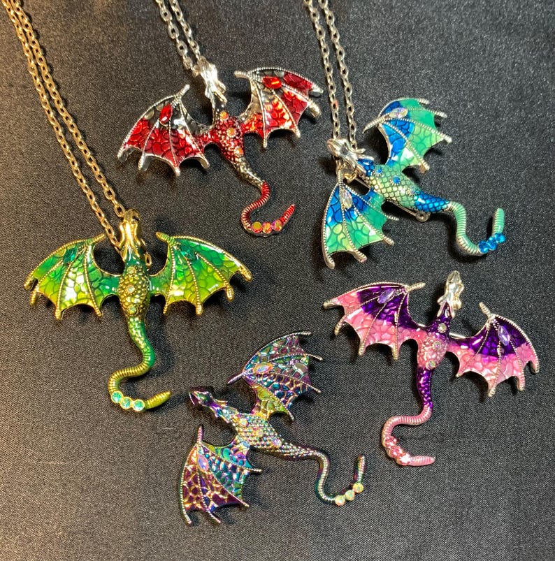 May include: Five dragon-shaped pendants on silver-toned chains. The dragons feature colorful enamel wings and bodies in red, green, blue, purple, and iridescent patterns, with small embedded crystals along their tails and bodies.
