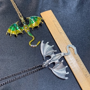 May include: Two dragon pendants on chains next to a wooden ruler. One dragon is gold-toned with green and yellow enamel wings and small clear stones. The other is a silver-toned metal dragon. The ruler shows the pendants are about 3 inches long.