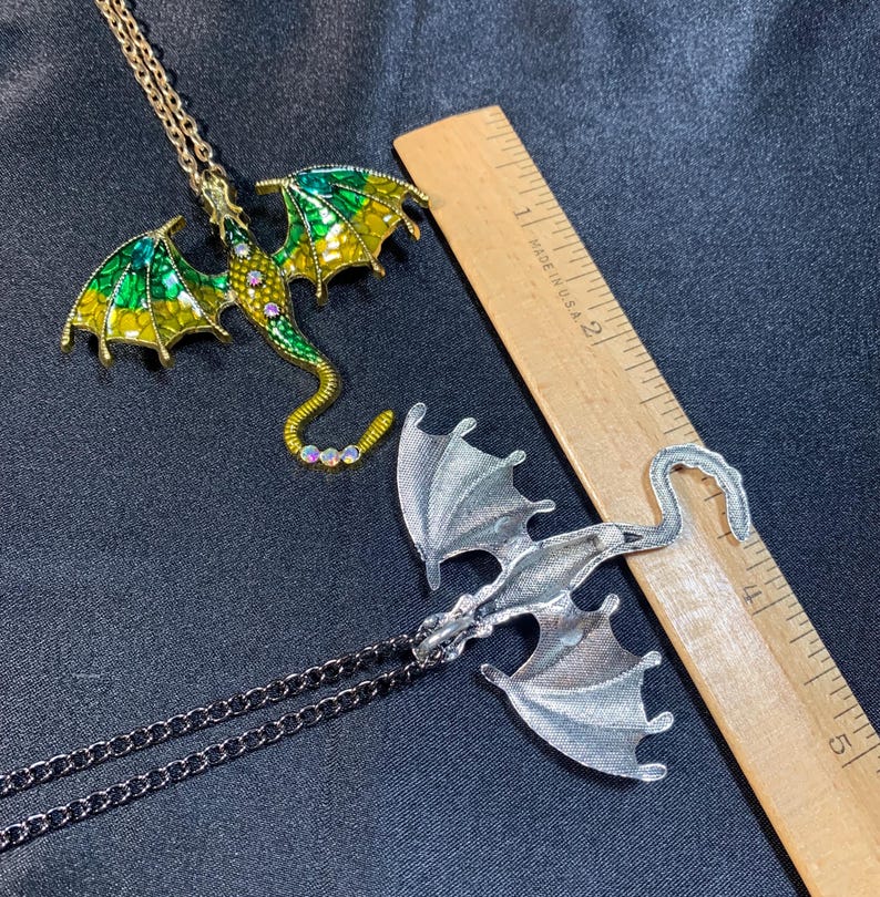 May include: Two dragon pendants on chains next to a wooden ruler. One dragon is gold-toned with green and yellow enamel wings and small clear stones. The other is a silver-toned metal dragon. The ruler shows the pendants are about 3 inches long.