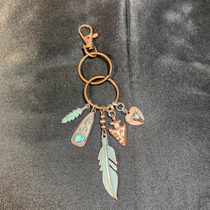 May include: A copper-toned key chain with multiple charms, including a turquoise heart, a feather, and a small arrowhead. The charms are attached to a double ring and a lobster clasp.