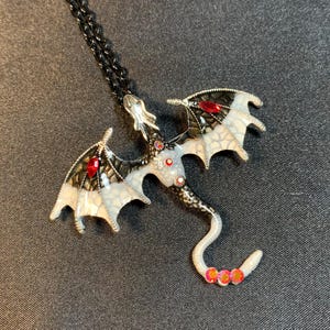 May include: A dragon pendant necklace featuring a black metal chain and a dragon-shaped charm. The dragon has white and black wings with red gemstone accents, a white body with small red stones, and a curved tail tipped with three red stones.