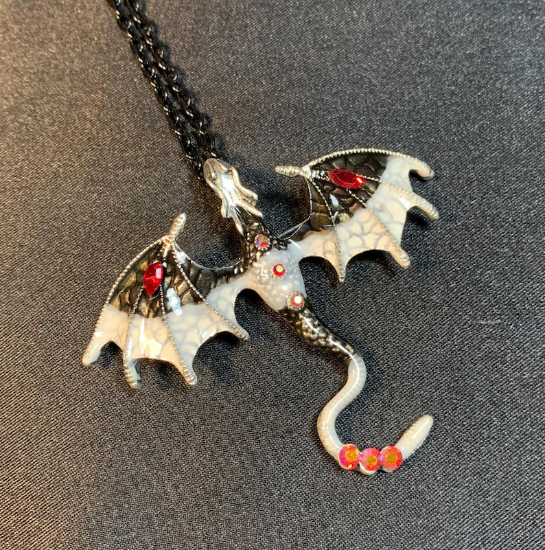 May include: A dragon pendant necklace featuring a black metal chain and a dragon-shaped charm. The dragon has white and black wings with red gemstone accents, a white body with small red stones, and a curved tail tipped with three red stones.