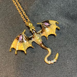 May include: A gold-toned metal dragon pendant necklace featuring enamel wings with a yellow and black scale pattern, accented by marquise-cut purple stones on each wing, small clear rhinestones on the body, and iridescent stones on the tail.