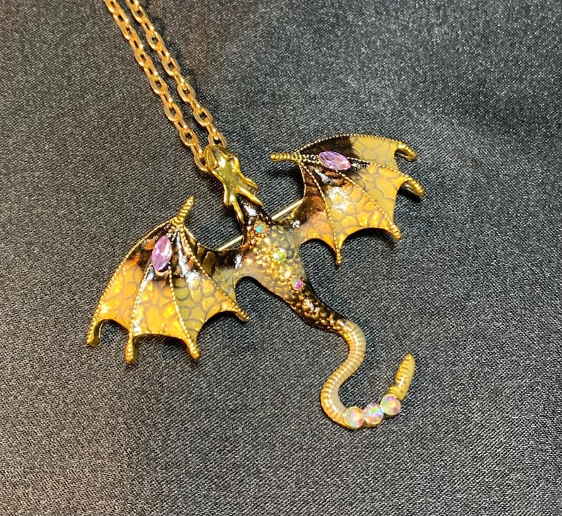 May include: A gold-toned metal dragon pendant necklace featuring enamel wings with a yellow and black scale pattern, accented by marquise-cut purple stones on each wing, small clear rhinestones on the body, and iridescent stones on the tail.