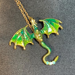 May include: A gold-toned metal dragon pendant necklace featuring vibrant green and yellow enamel wings with a textured scale pattern. The dragon body is adorned with small clear crystals and three light blue stones at the tip of its curved tail.