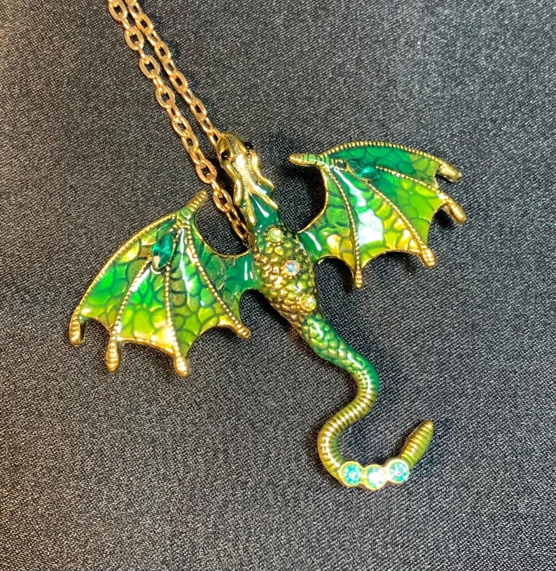 May include: A gold-toned metal dragon pendant necklace featuring vibrant green and yellow enamel wings with a textured scale pattern. The dragon body is adorned with small clear crystals and three light blue stones at the tip of its curved tail.