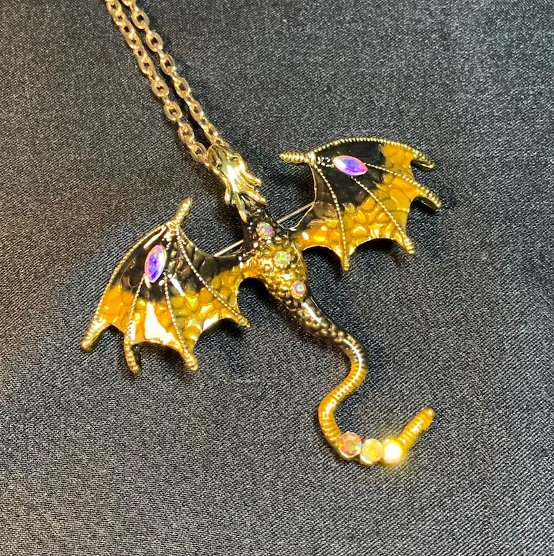 May include: A gold-toned dragon pendant necklace featuring black and yellow enamel wings with iridescent marquise-cut stones. The dragon body is textured with small gold bumps and accented with tiny clear rhinestones along the back and tail.