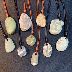 May include: A collection of eight necklaces featuring smooth, natural river stones in various shades of gray, green, and tan. Each stone is suspended from a simple cord, with some stones featuring white mineral veins or speckled patterns.