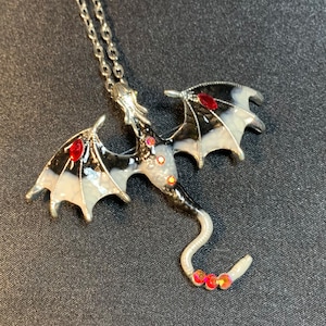 May include: A silver-toned metal dragon pendant necklace featuring black and white enamel detailing on the wings and body, accented with small, round, iridescent red rhinestones embedded along the wings, torso, and the tip of the curved tail.