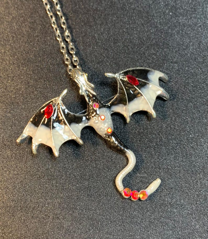 May include: A silver-toned metal dragon pendant necklace featuring black and white enamel detailing on the wings and body, accented with small, round, iridescent red rhinestones embedded along the wings, torso, and the tip of the curved tail.