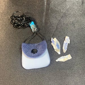 May include: A small blue leather bag with a black button closure hangs from a black wire necklace with blue and turquoise beads. Three small blue and white crystal stones are on a dark gray surface.