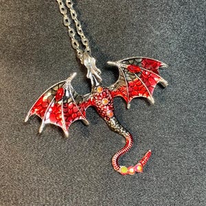 May include: A silver-toned metal dragon pendant necklace featuring red and black enamel detailing on the wings and body. The dragon design includes small iridescent circular accents along the neck and tail, suspended from a matching silver-toned chain.