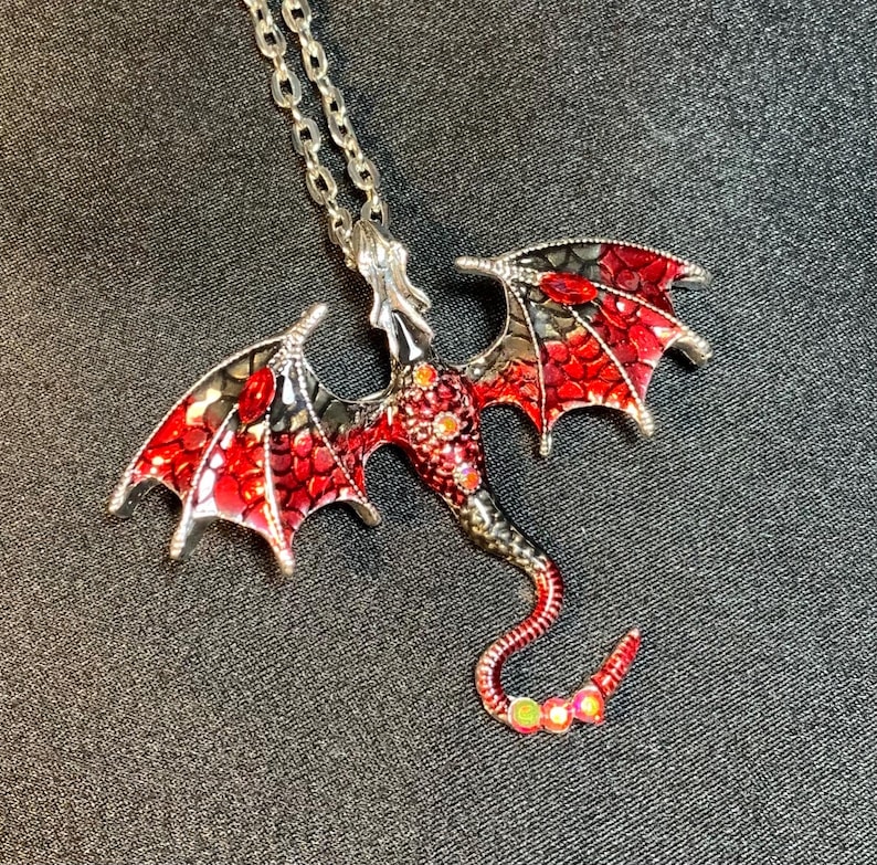 May include: A silver-toned metal dragon pendant necklace featuring red and black enamel detailing on the wings and body. The dragon design includes small iridescent circular accents along the neck and tail, suspended from a matching silver-toned chain.