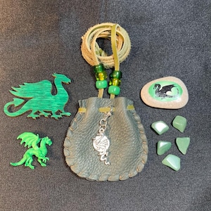 May include: A dark green leather drawstring pouch with a silver dragon charm, a green wooden dragon cutout, a small green plastic dragon figure, a stone with a black dragon print, and five small green tumbled stones with a leather cord necklace.