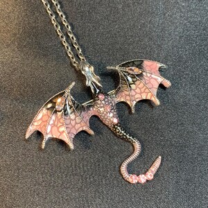 May include: A silver-toned metal dragon pendant necklace featuring enamel wings in shades of pink and black with a scale-like pattern. The dragon body is accented with small pink crystals embedded along the back and the curved tail.