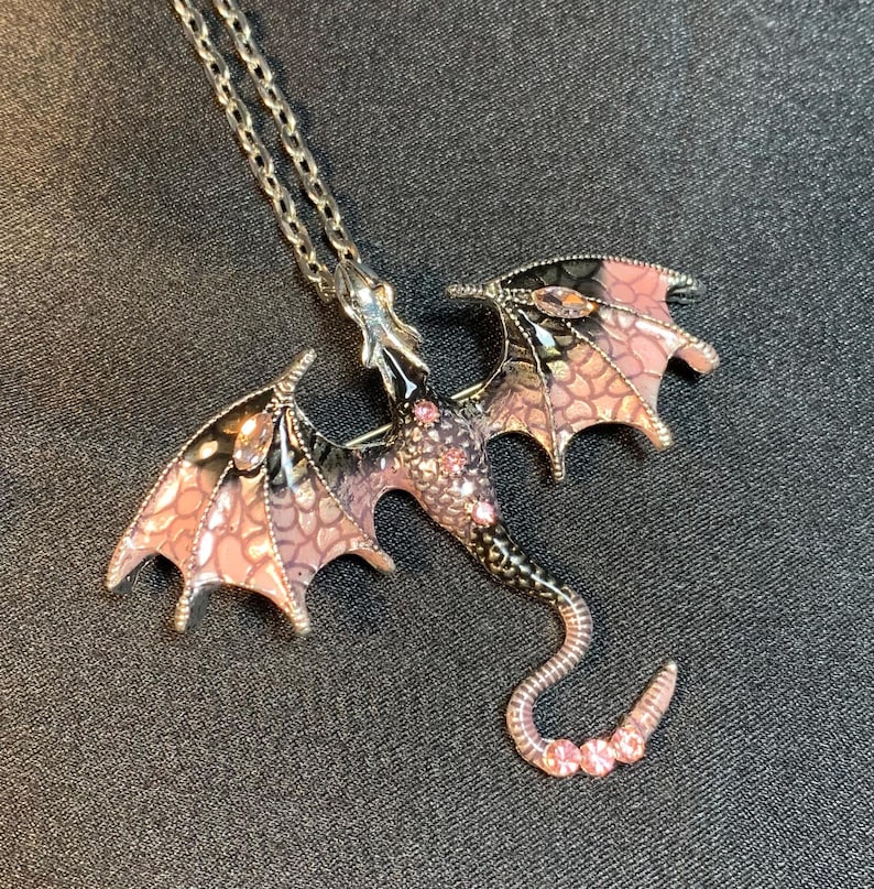 May include: A silver-toned metal dragon pendant necklace featuring enamel wings in shades of pink and black with a scale-like pattern. The dragon body is accented with small pink crystals embedded along the back and the curved tail.
