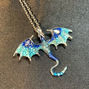 May include: A silver-toned metal dragon pendant necklace featuring enamel wings in shades of blue and teal with a scale pattern. The dragon body is embellished with small blue crystals and iridescent stones on the wings and a curved tail.