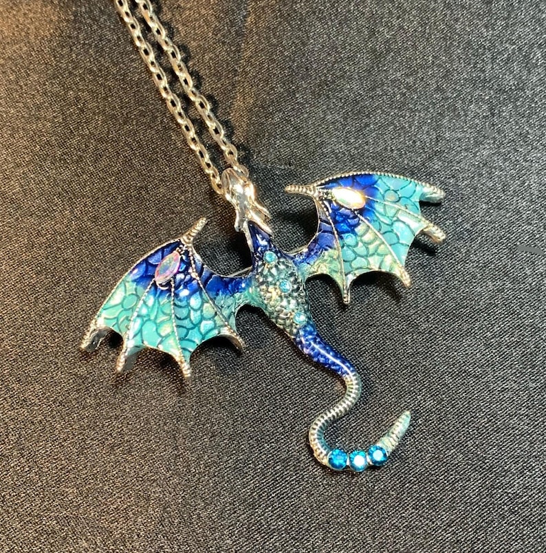 May include: A silver-toned metal dragon pendant necklace featuring enamel wings in shades of blue and teal with a scale pattern. The dragon body is embellished with small blue crystals and iridescent stones on the wings and a curved tail.