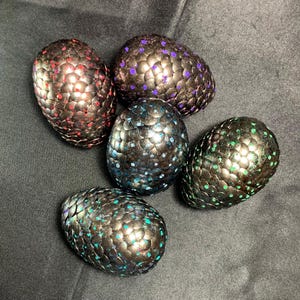 May include: Five decorative dragon eggs with textured, scale-like surfaces. The eggs are painted in various colors, including red, purple, blue, and green, with a metallic sheen. The eggs are arranged on a dark gray surface.