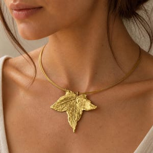 Può includere: Una collana color oro con un ciondolo a forma di foglia strutturata. Il design della foglia presenta venature dettagliate ed è attaccato a un sottile girocollo a più fili. La collana è indossata su uno sfondo neutro.