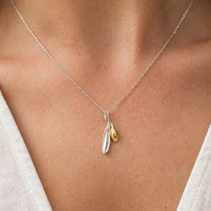 May include: A silver necklace with a pendant featuring two leaf-shaped charms, one silver and one gold. The necklace is worn against a white top.