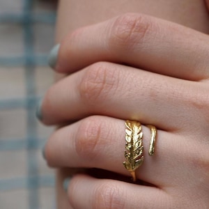 Puede incluir: Un anillo dorado con diseño de hoja. El anillo se envuelve alrededor del dedo, con una hoja detallada en un lado y una banda lisa y curvada en el otro. El anillo se lleva en una mano con esmalte de uñas azul claro.